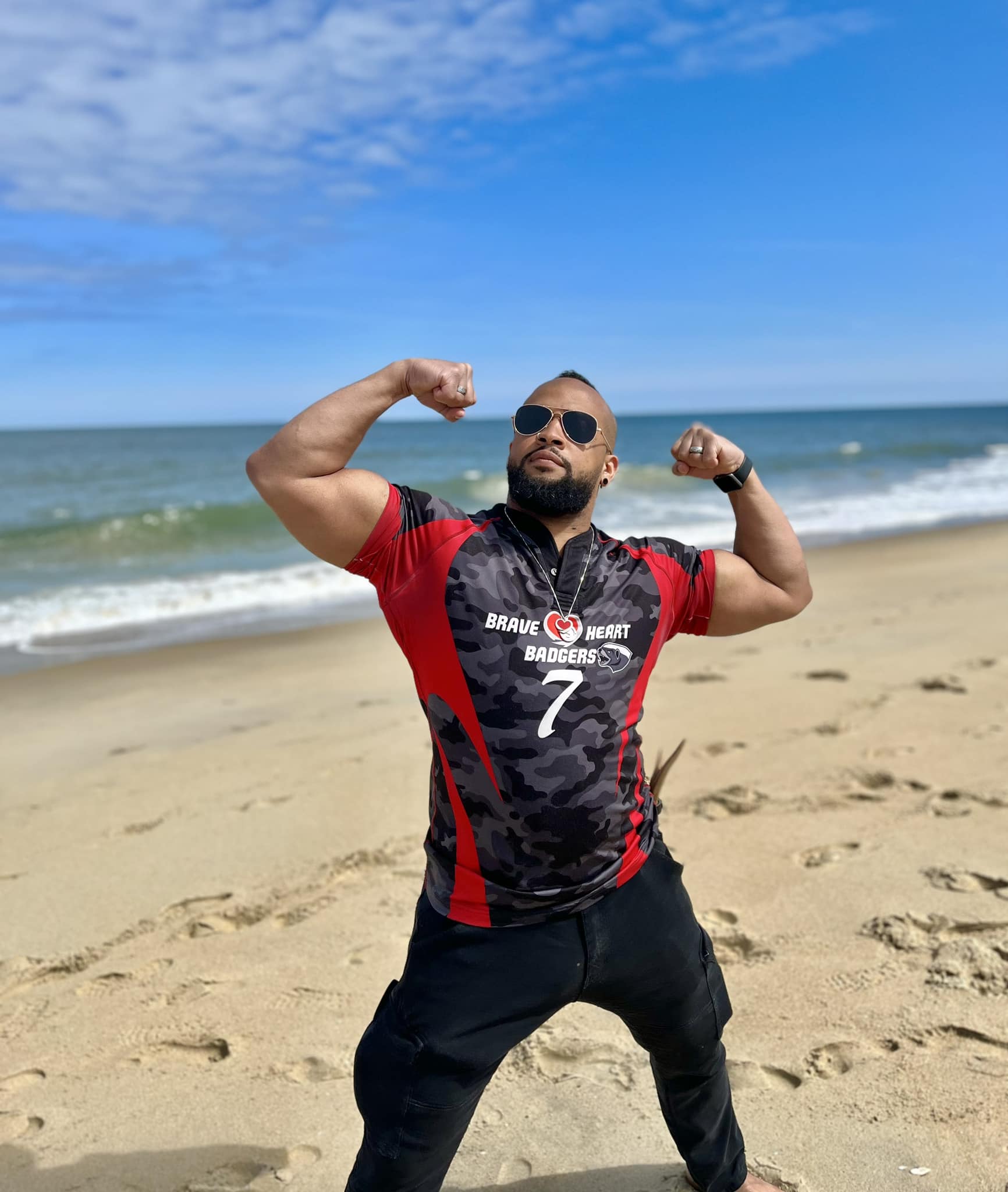 Coach Nick Friday stands with legs extended and arms in a full flex in his Braveheart Badgers shirt on a shoreline. "Be Braveheart" is written on top of the image.