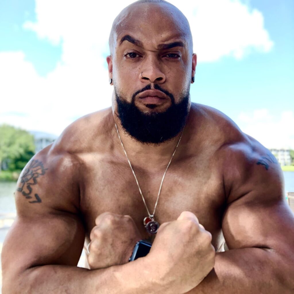 Close up of Coach Nick standing shirtless in front of a lake with two clenched fists in a firm flex.