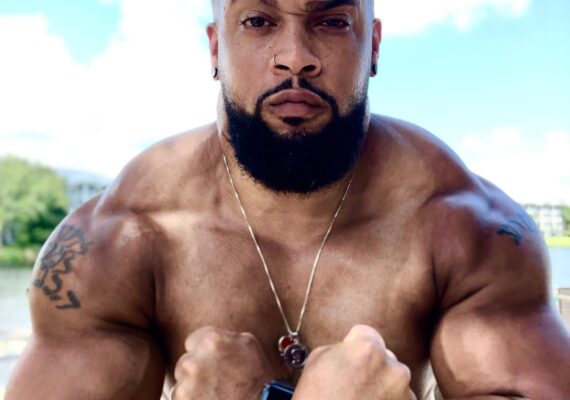 Close up of Coach Nick standing shirtless in front of a lake with two clenched fists in a firm flex.